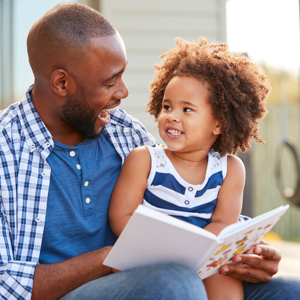 Young girl sitting on dad's knee reading a book together and laughing.