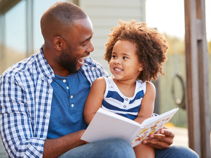 Young girl sitting on dad's knee reading a book together and laughing.