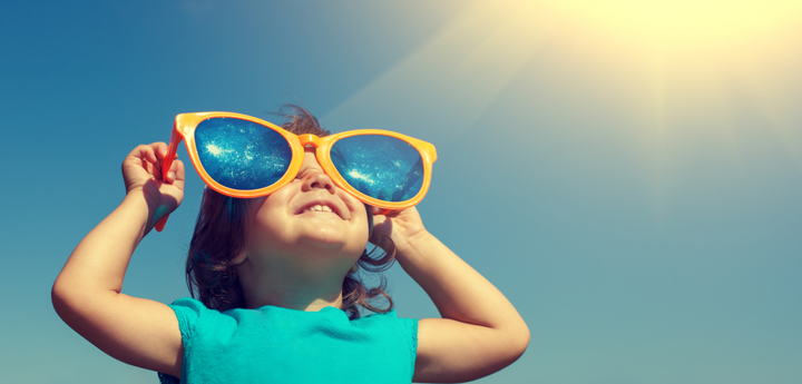 Young girl smiling whilst wearing oversized yellow sunglasses looking up at the sky.
