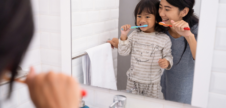 Mother and young girl standing in front of bathroom sink brushing their teeth and looking in the mirror.