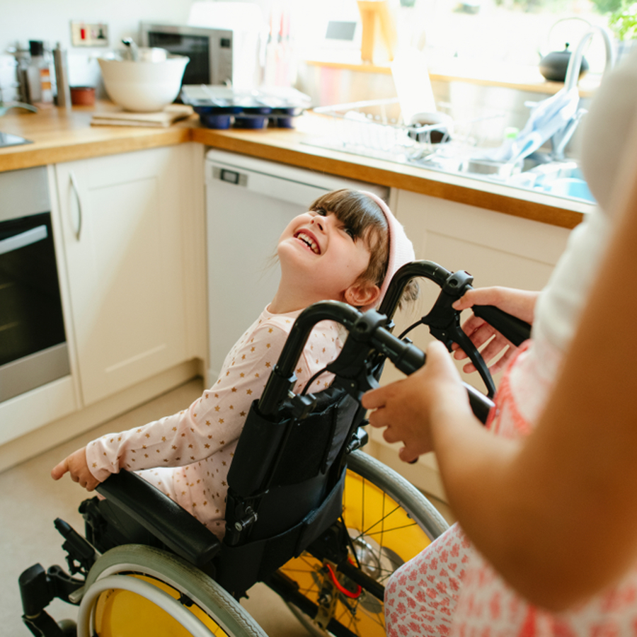 Young girl in a wheelchair smiling up at an adult standing behind her and pushing her wheelchair. Situated in a kitchen