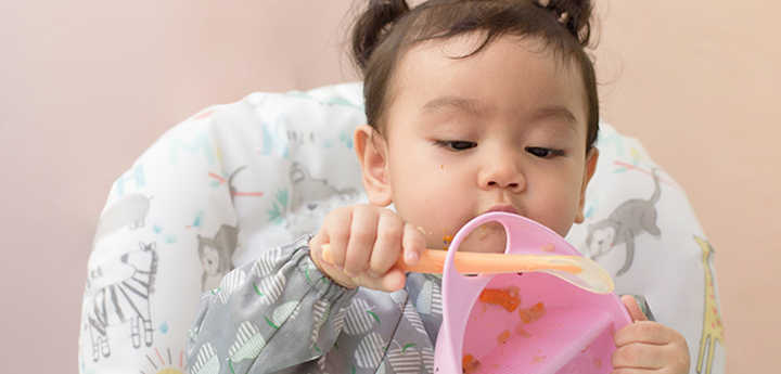 Baby in high chair playing with pink bowl with orange spoon