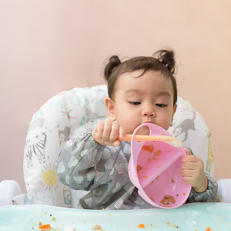 Baby in high chair playing with pink bowl with orange spoon