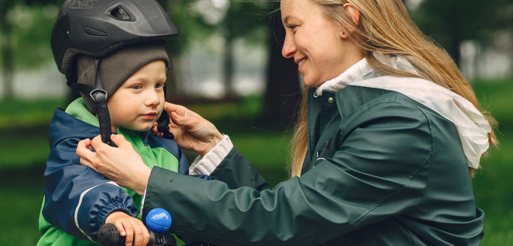 Toddler boy sitting on balance bike in a rain suit. Mum kneeling down next to him strapping his helmet to his head