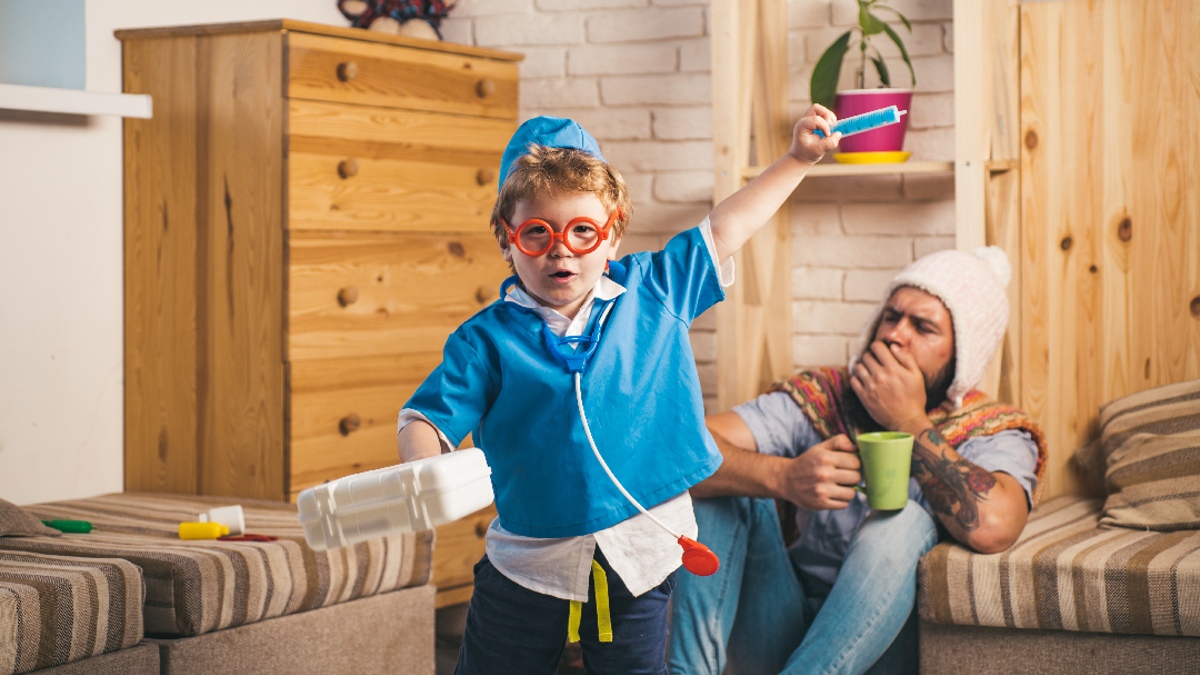 Child in nurses outfit with a toy stethoscope, toy syringe and a carrier box. Adult man in the background coughing and sat on the ground with a woolly hat and green mug 