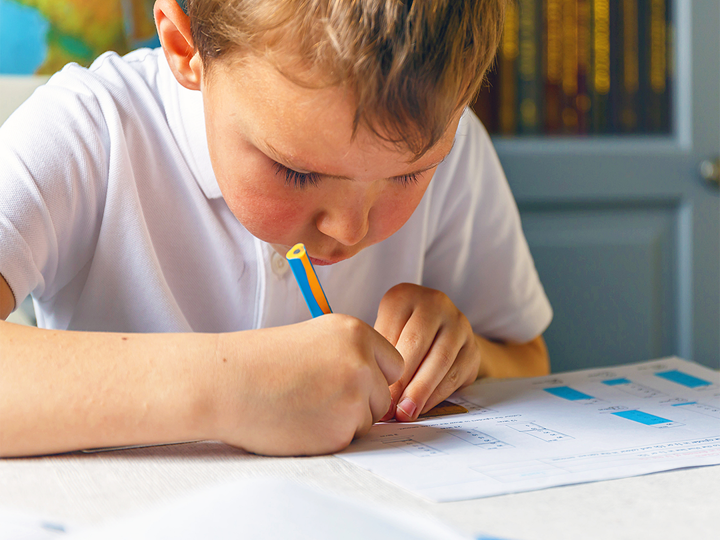 Young boy dressed in school uniform sitting in a classroom at a desk writing on a piece of paper