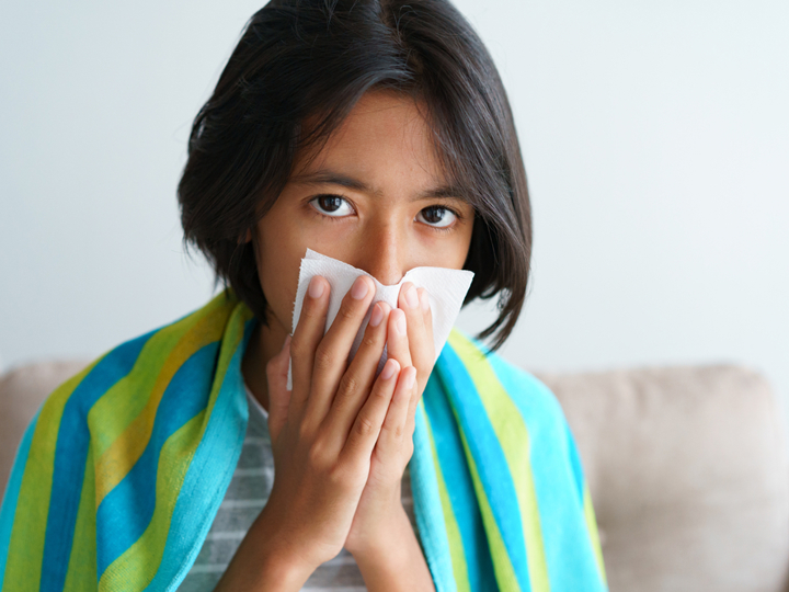 Girl sitting on sofa wrapped in a blanket, blowing nose into a tissue.