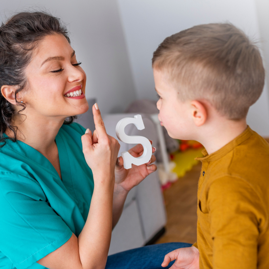 Woman pointing to lips whilst pronouncing the letter 's' and holding a letter 's' with a young boy standing in front of her