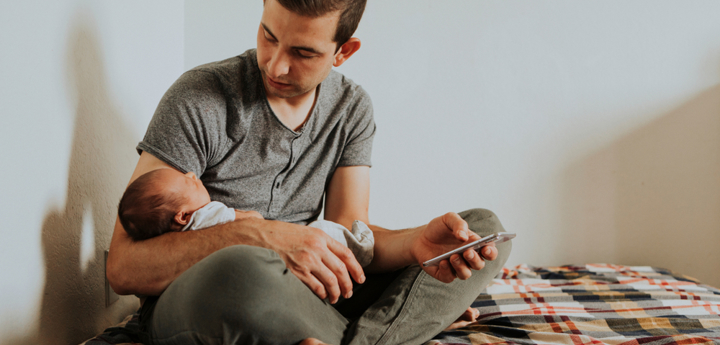 Dad sitting on bed looking down at his baby which he is holding in his arm. His other hand is holding his phone.