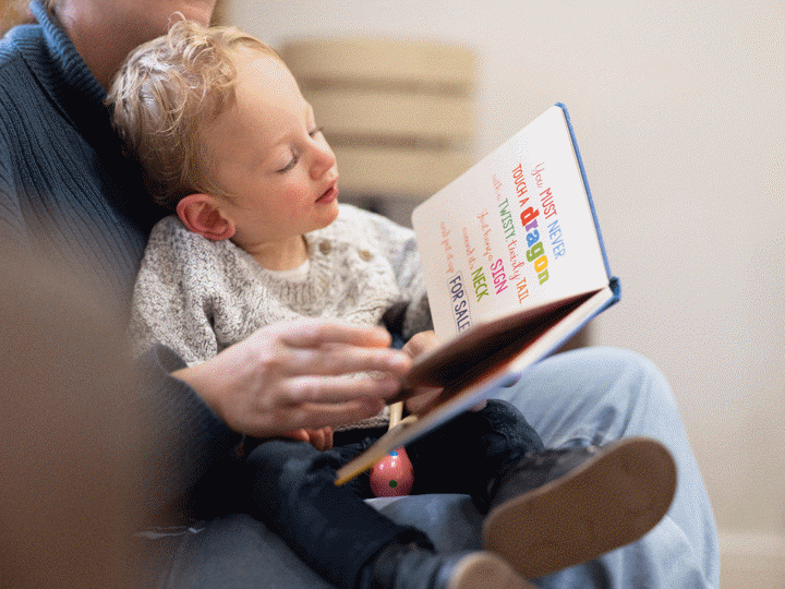 Boy looking at book whilst sitting in his parents lap. The parent is holding the book reading it to the young boy.