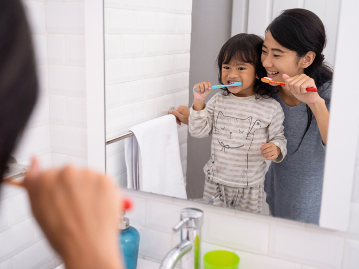 Young girl brushing their teeth with their mum looking into the bathroom mirror