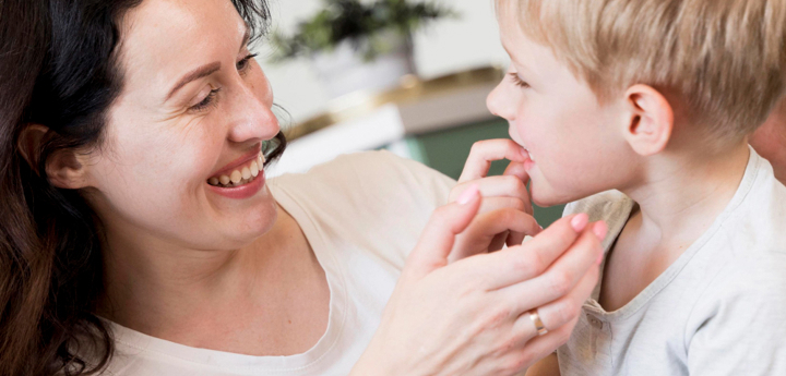 Young boy smiling at his mum, holding his hand to his mouth. The mum is smiling back at him and reaching towards his hand