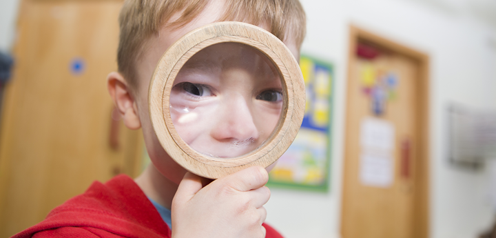blonde haired young boy looking through wooden magnifying glass, his eyes and nose are exaggerated. background is a blurred cream wall with 2 wooden doors