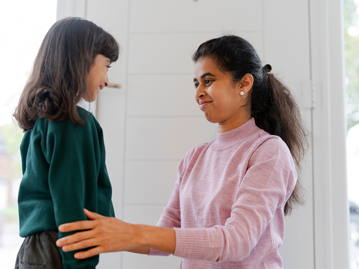 Adult kneeling in front of girl dressed in school uniform. Adult lightly touching child's arms, both smiling.