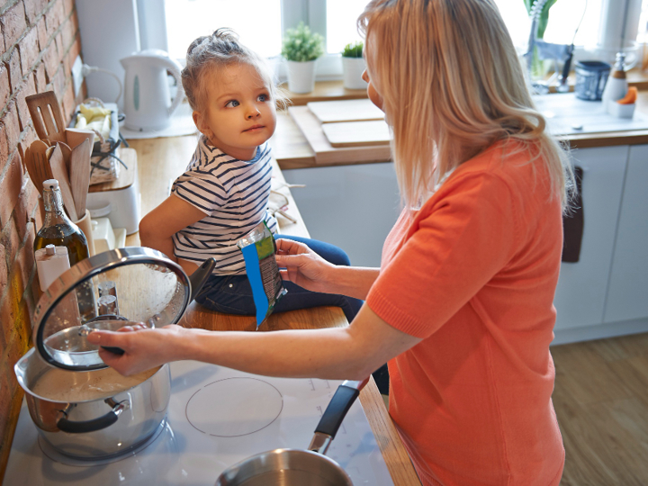 Young girl sitting on kitchen worktop looking at adult woman who is cooking on stove